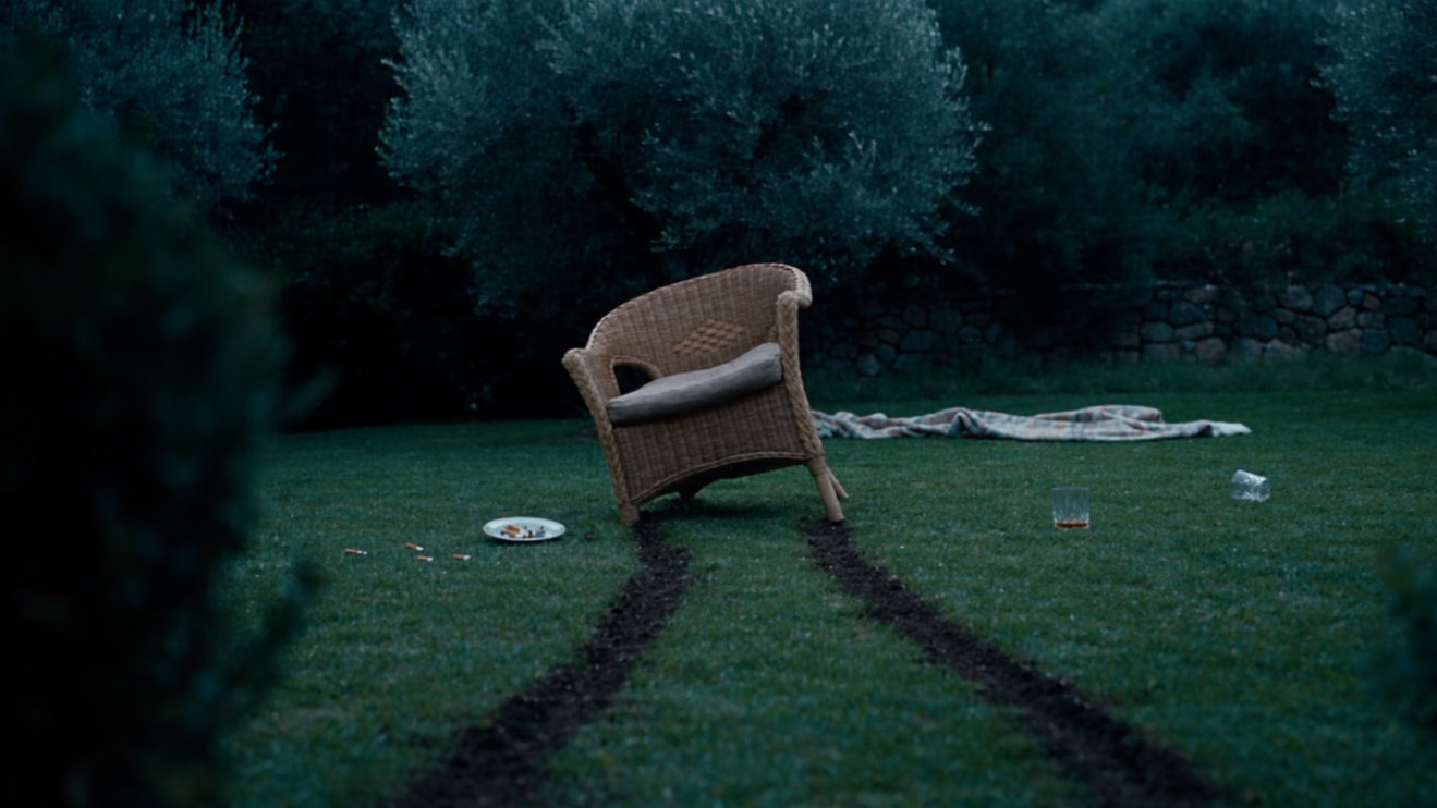 A wicker chair on a lawn at dusk, two furrows of torn earth running back from its legs