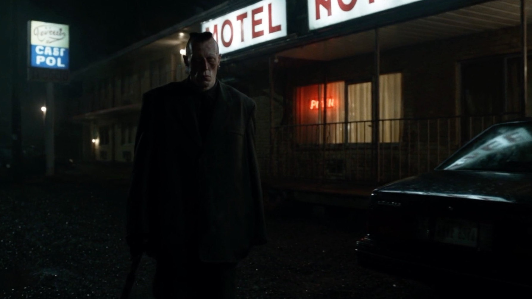 A gaunt man in a long coat standing in the rain outside a motel at night
