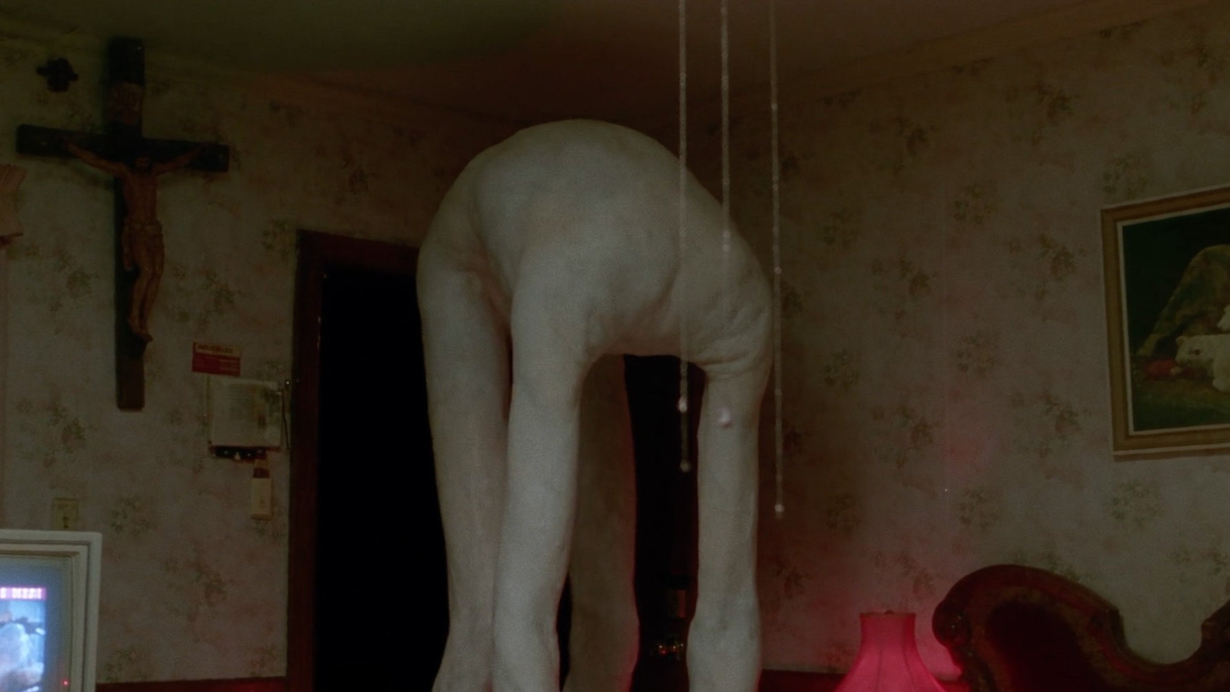 A pale, headless figure bent double in a small wallpapered room under a crucifix