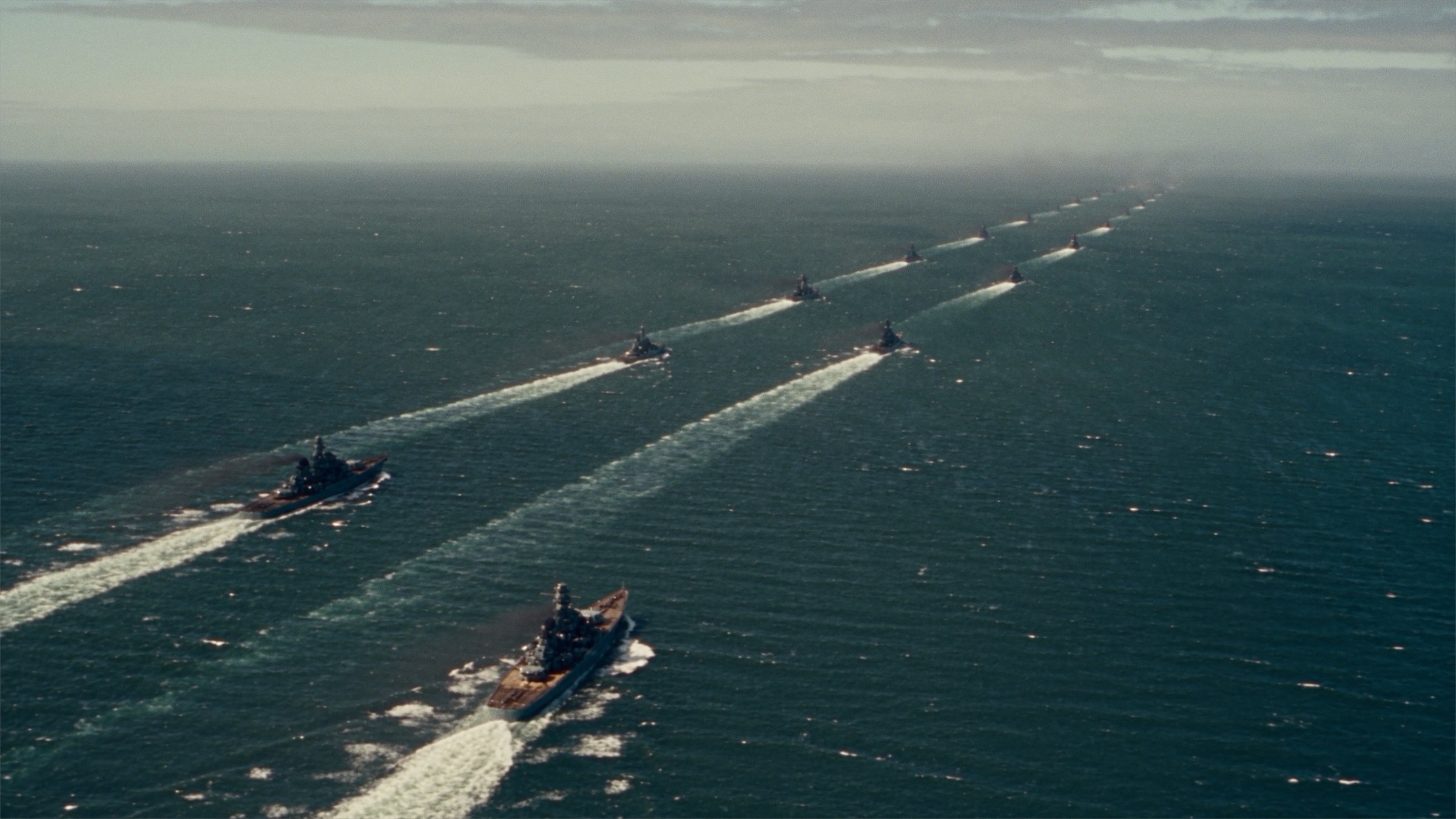 A fleet of warships in two long columns cutting white wakes across open sea, seen from high above