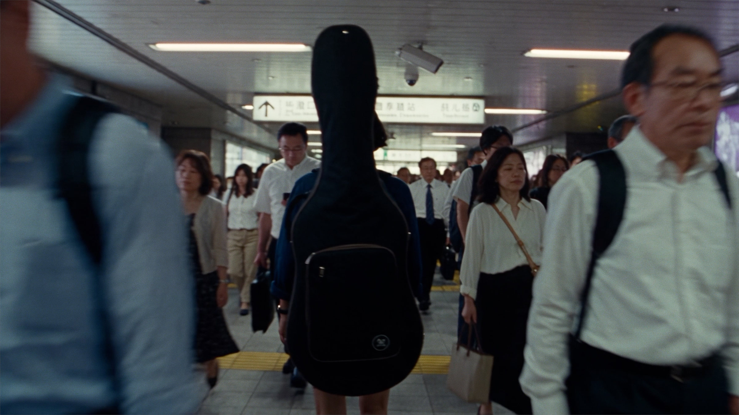 A musician with a guitar case on her back, walking against a rush-hour crowd in a station underpass